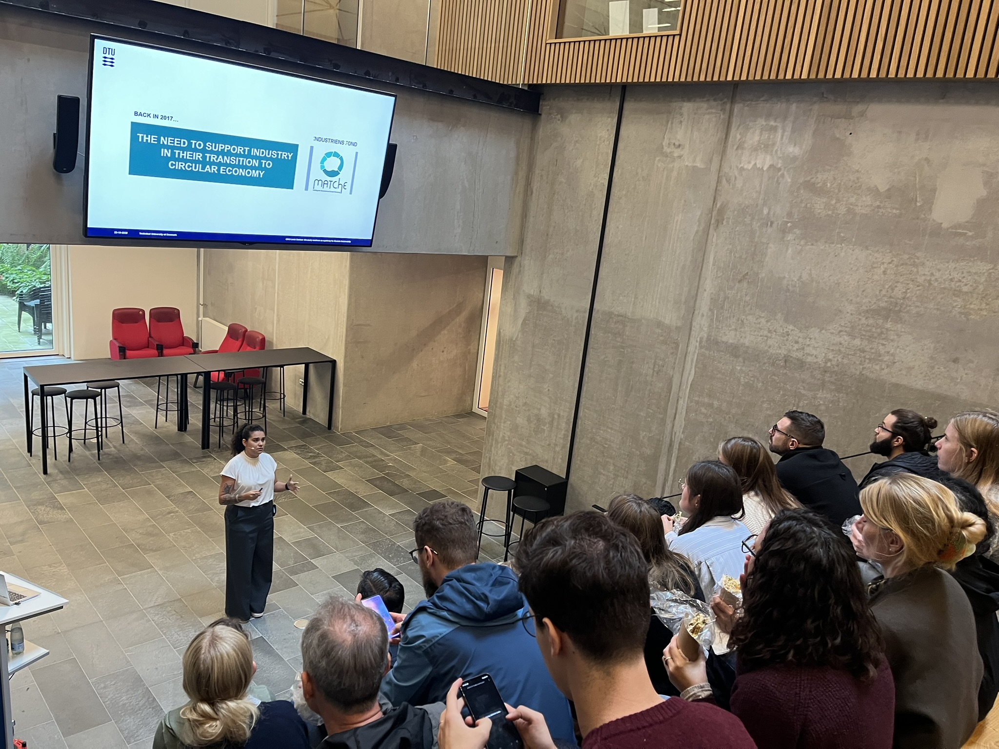 A woman presents to an audience with a screen that says "The need to support industry in their transition to circular economy"