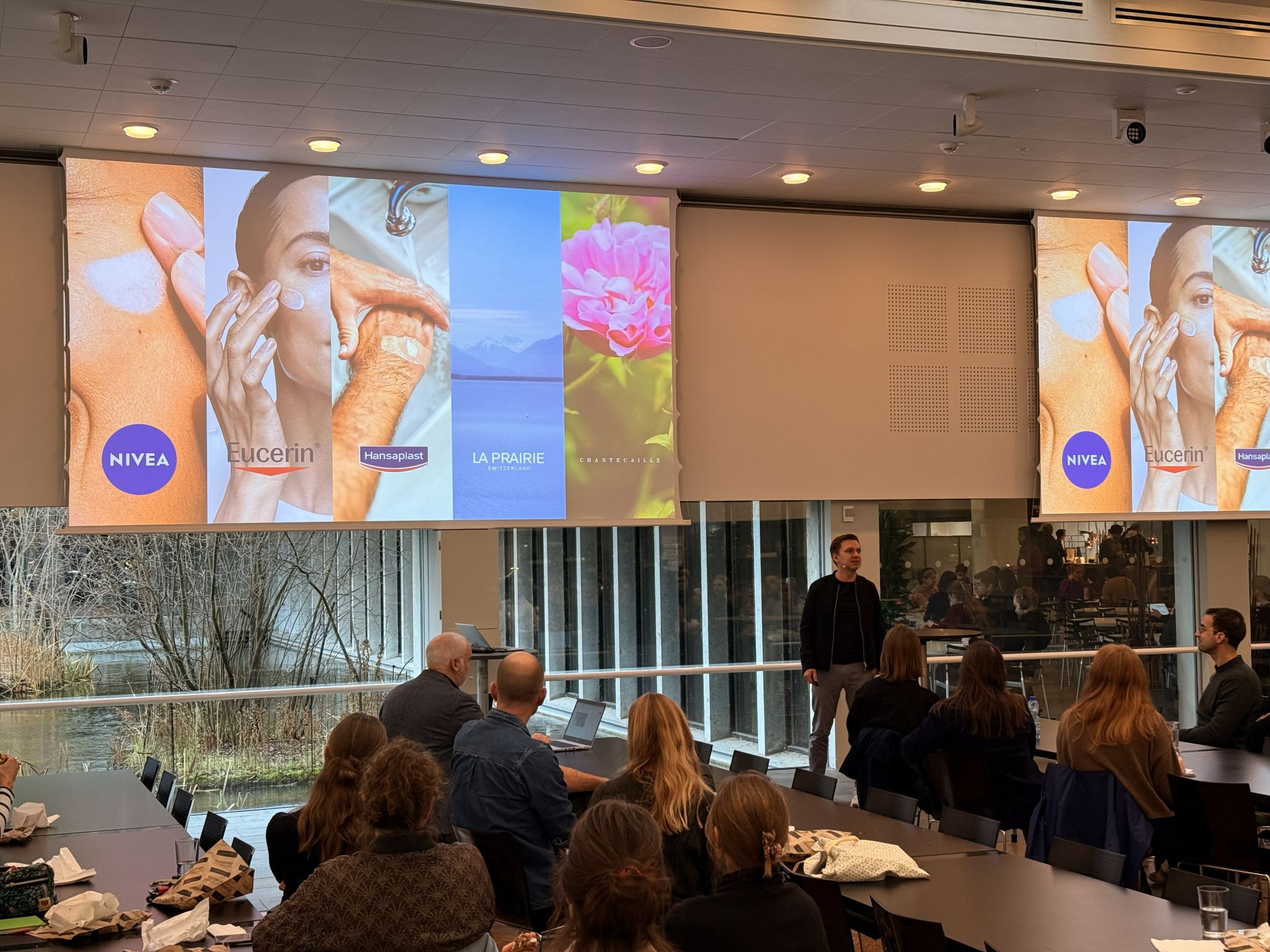 A main is presenting in a busy hall in front of a screen showing beauty products